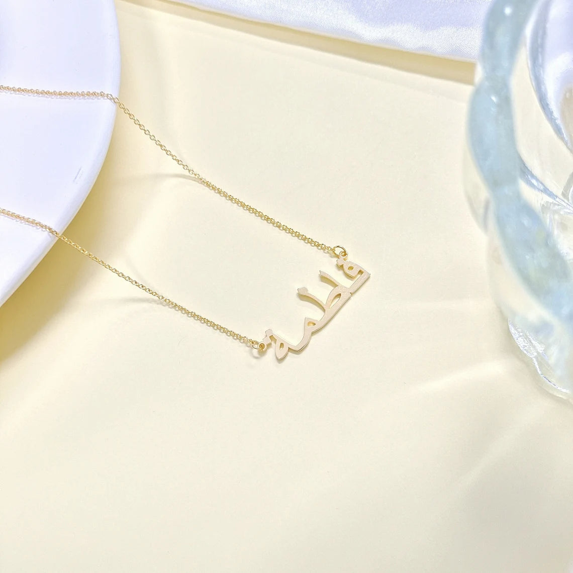 Custom Arabic name necklace in gold, elegantly displayed on a beige surface, showcasing personalized jewelry design for stylish and unique fashion accessories.