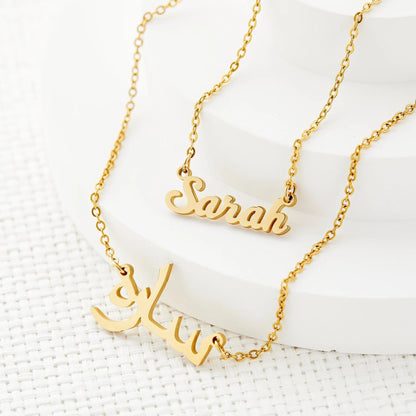 Close-up of two 18k gold plated custom name necklaces by Thaheb, one with "Sarah" in English and the other with a name in Arabic script.