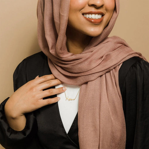 A woman in a pink hijab and black top smiles while showcasing an 18k gold plated custom Arabic name necklace by Thaheb.