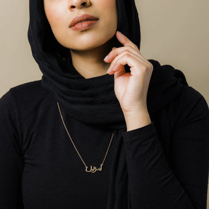 A woman wearing a black hijab and black top showcases a beautiful 18k gold plated custom Arabic name necklace by Thaheb.