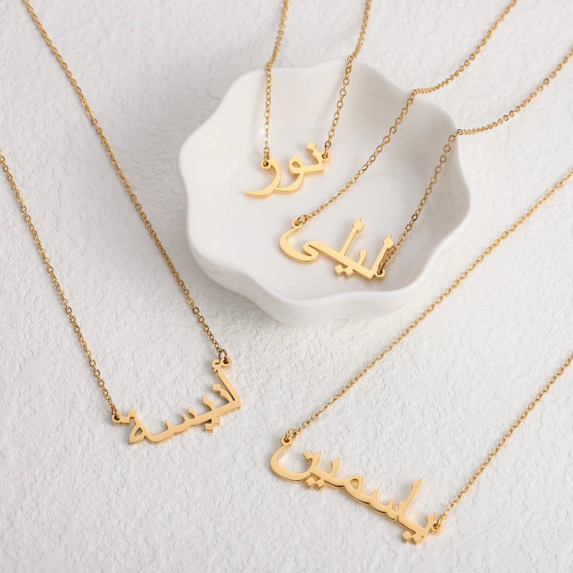 Collection of custom Arabic name necklaces in gold, with some necklaces elegantly displayed in a white dish and others laid out on a white surface, showcasing personalized jewelry design for unique fashion accessories.